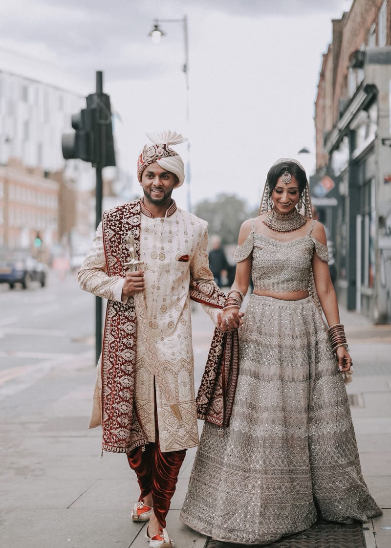 Couple walking hand in hand down a city street, the groom in a sherwani and turban, the bride in a silver lehenga