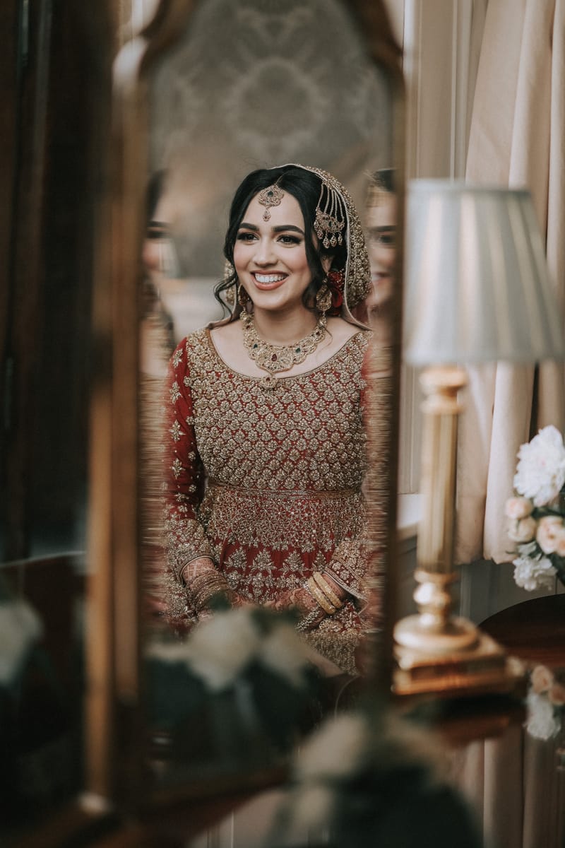 Smiling bride in red and gold reflected in an antique mirror beside a table lamp