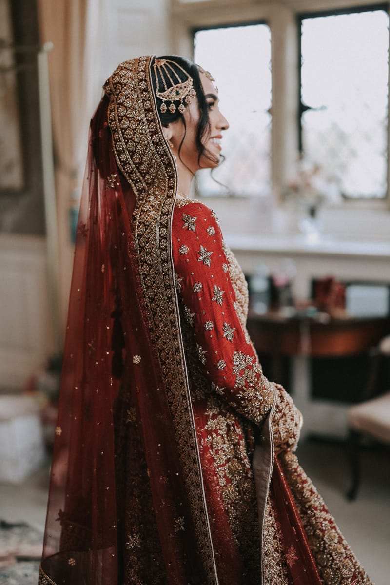 Bride in red with a heavy embroidered dupatta, standing in profile by a window