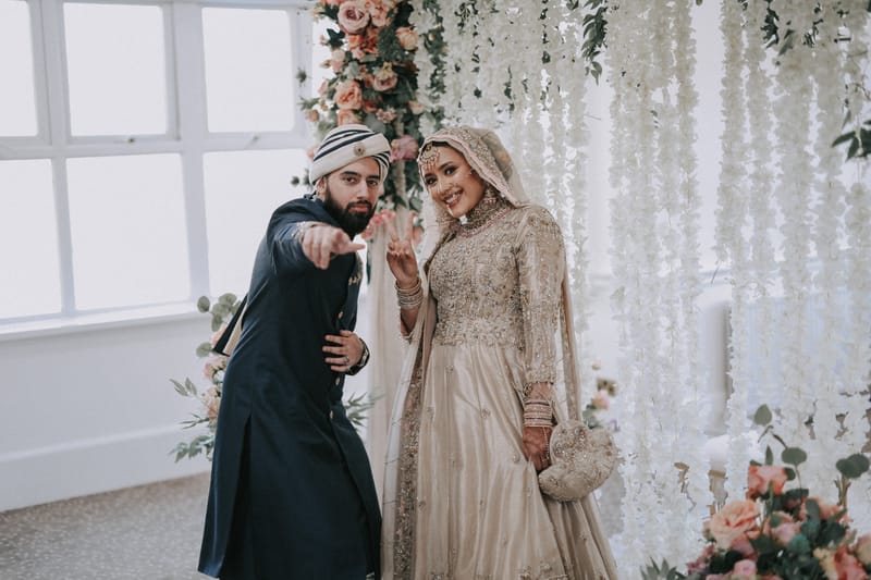 Groom in a turban striking a playful pose beside his smiling bride in front of a flower wall