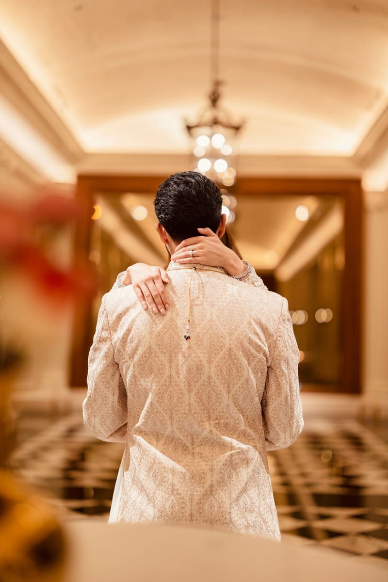 Bride's arms around the groom's neck in a warmly lit corridor, seen from behind the groom