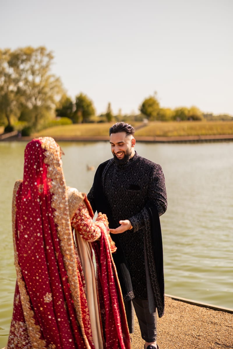 Groom smiling as he greets his bride in red beside a lake