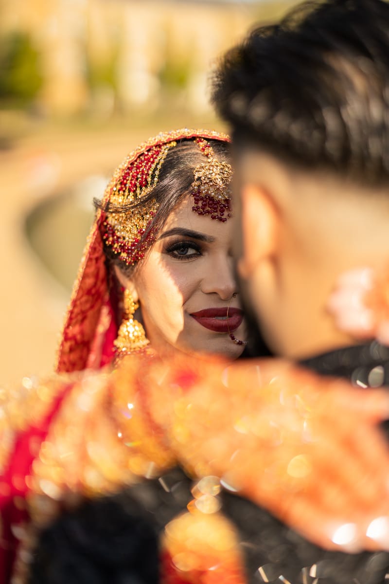 Bride in red with a gold tikka and jhumkas, smiling over the groom's shoulder