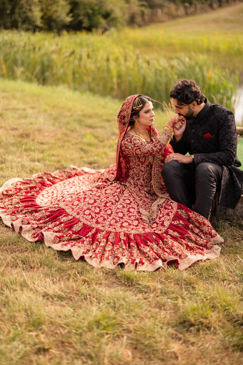 Groom kissing his bride's hand as they sit on the grass, her red lehenga spread around her