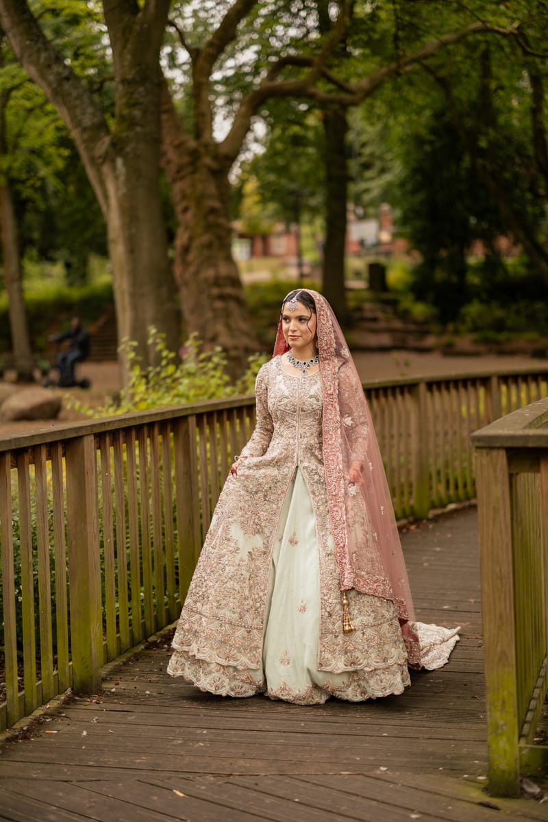 Bride in a blush pink lehenga walking across a wooden footbridge in a park