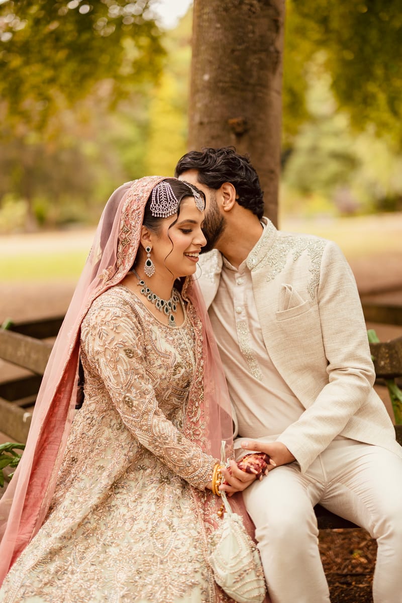 Groom resting his head against his smiling bride as they sit on a bench under a tree
