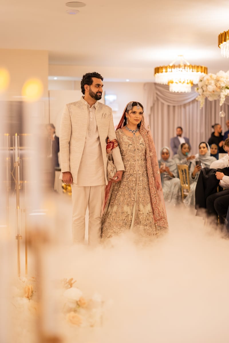 Couple making their entrance through low fog in a chandelier-lit hall as guests look on