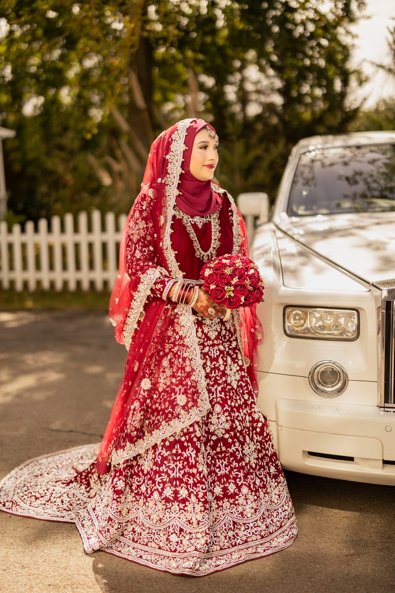 Bride in a red lehenga and hijab holding red roses beside a white wedding car