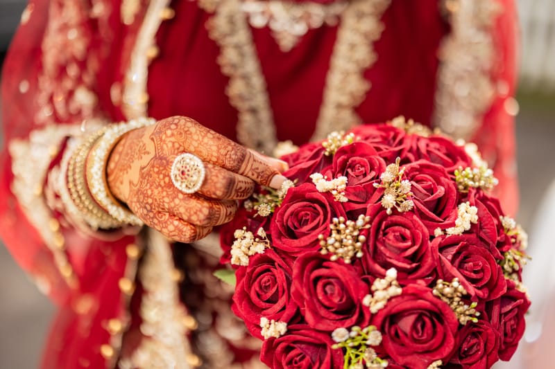 Close-up of a bride's Mehndi hand with gold bangles and a statement ring, holding red roses