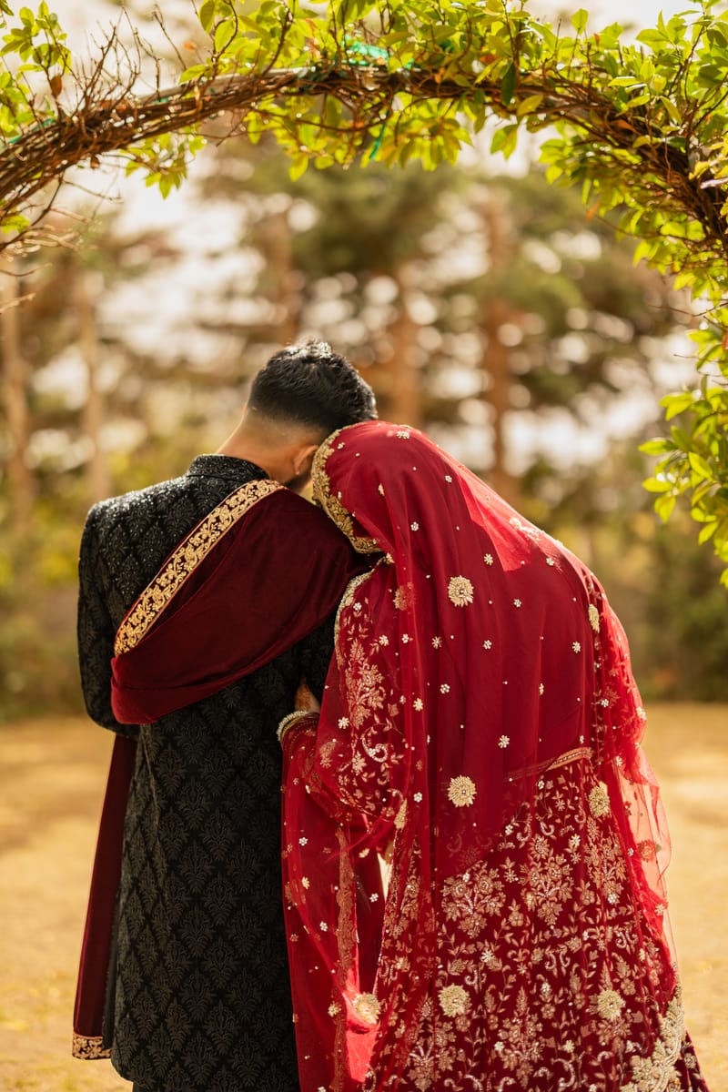 Bride in a red dupatta resting her head on the groom's shoulder under a leafy arch, seen from behind