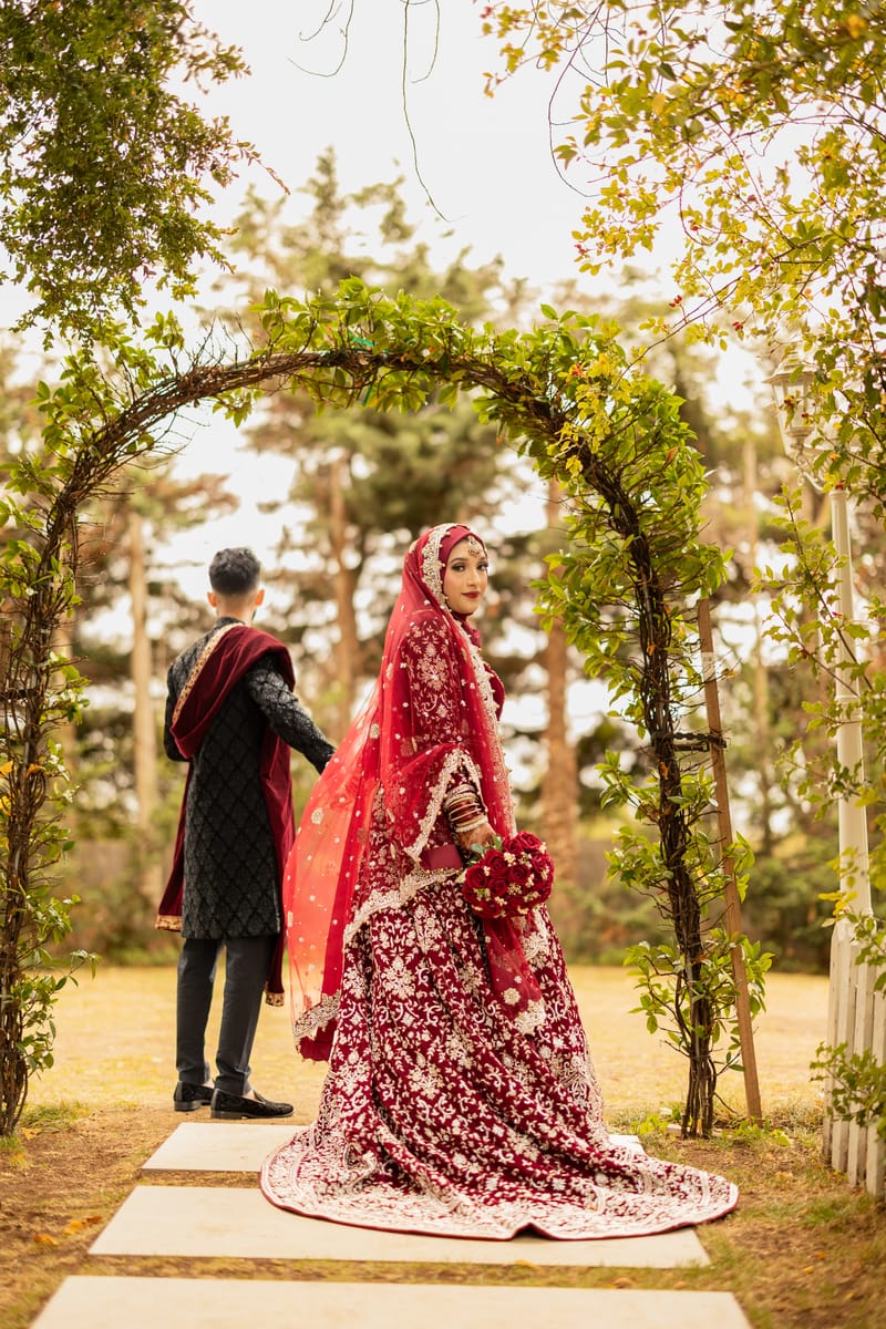 Bride in a red lehenga with a long train under a leafy arch, the groom waiting behind her
