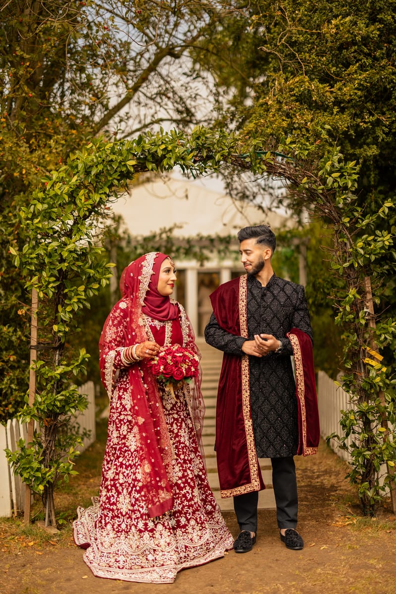 Bride in red and a hijab beside the groom in a black sherwani under a leafy archway