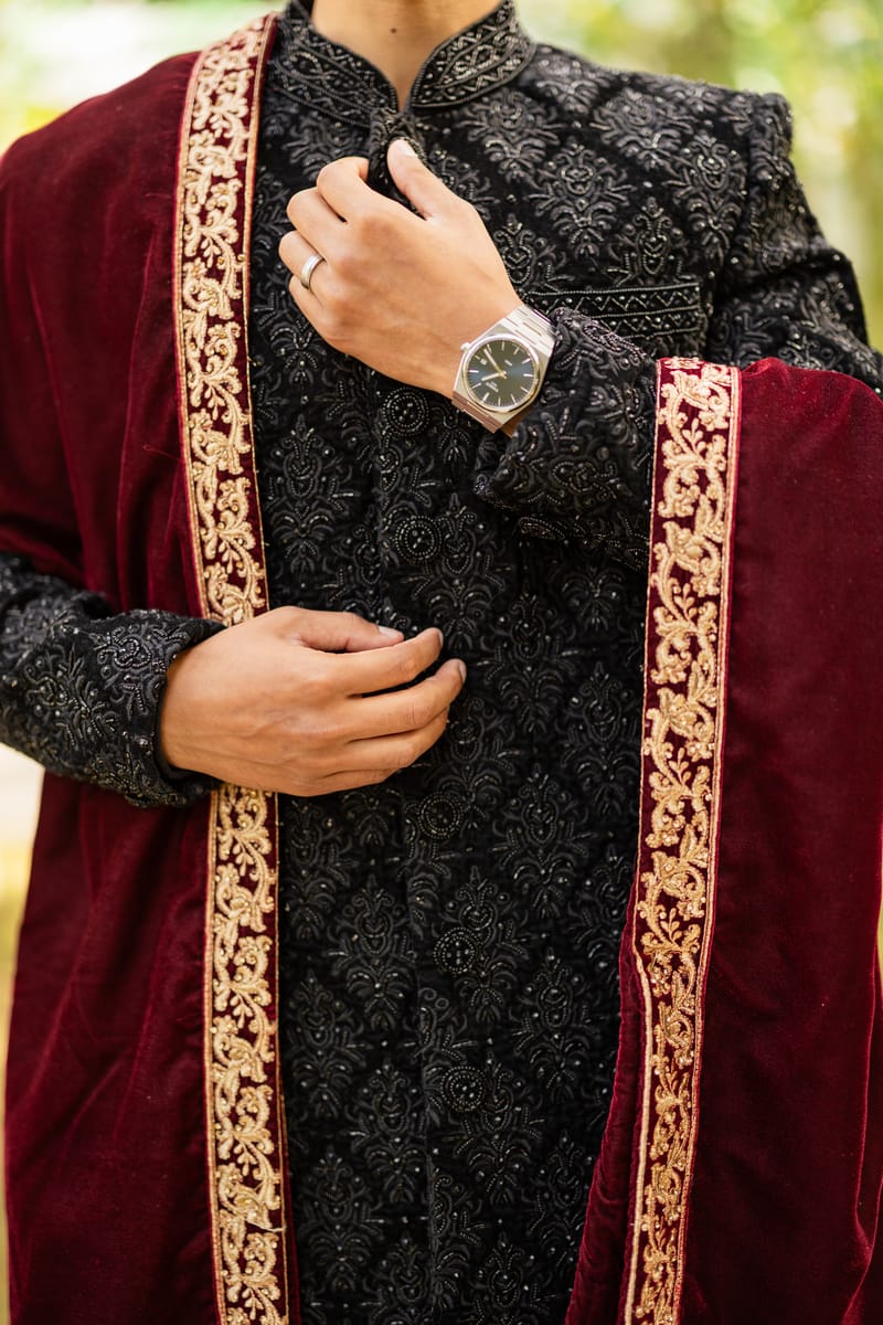 Close-up of the groom's black embroidered sherwani, watch and maroon velvet shawl with gold trim