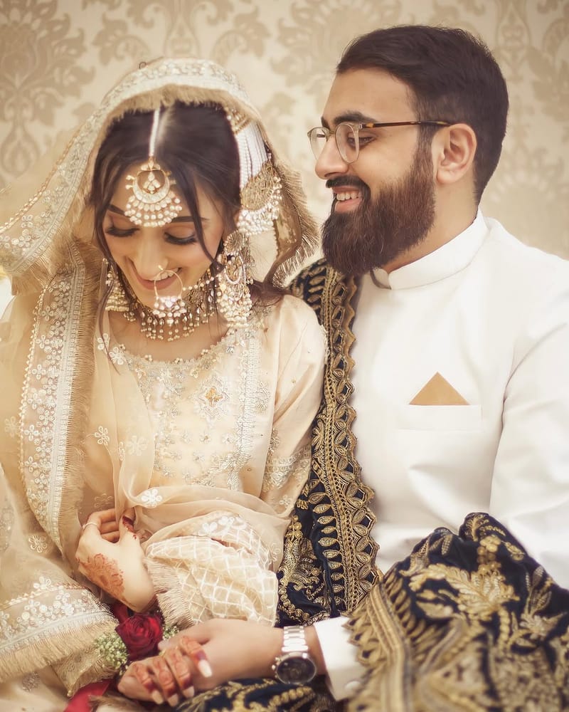 Laughing couple seated together, the bride in cream with a gold tikka and nath, the groom in a white sherwani